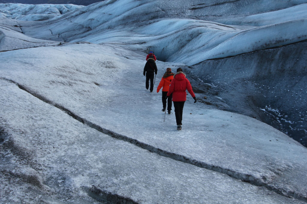 People in red jackets walking across a blue frozen glacier.