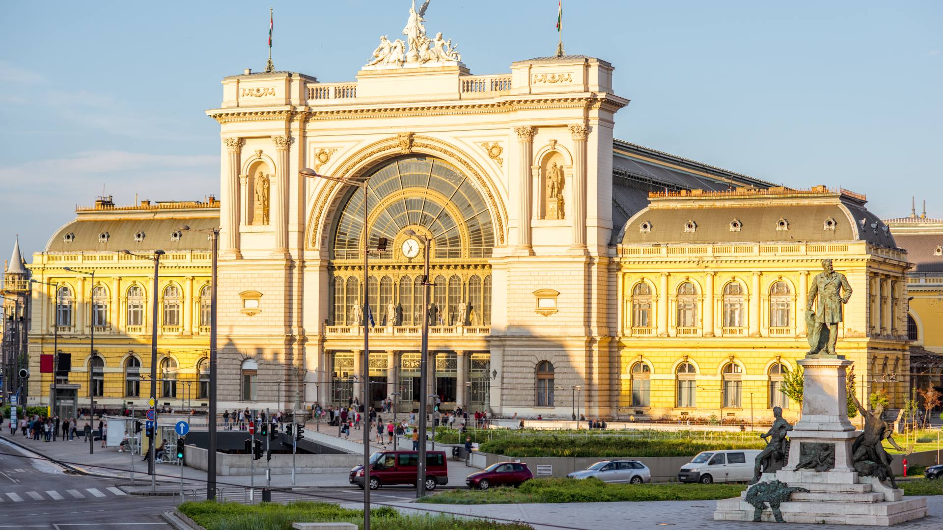 The grand neoclassical facade of Budapest Keleti railway station bathed in golden sunset light, with the Gábor Baross statue in the foreground and pedestrians gathered around the entrance.