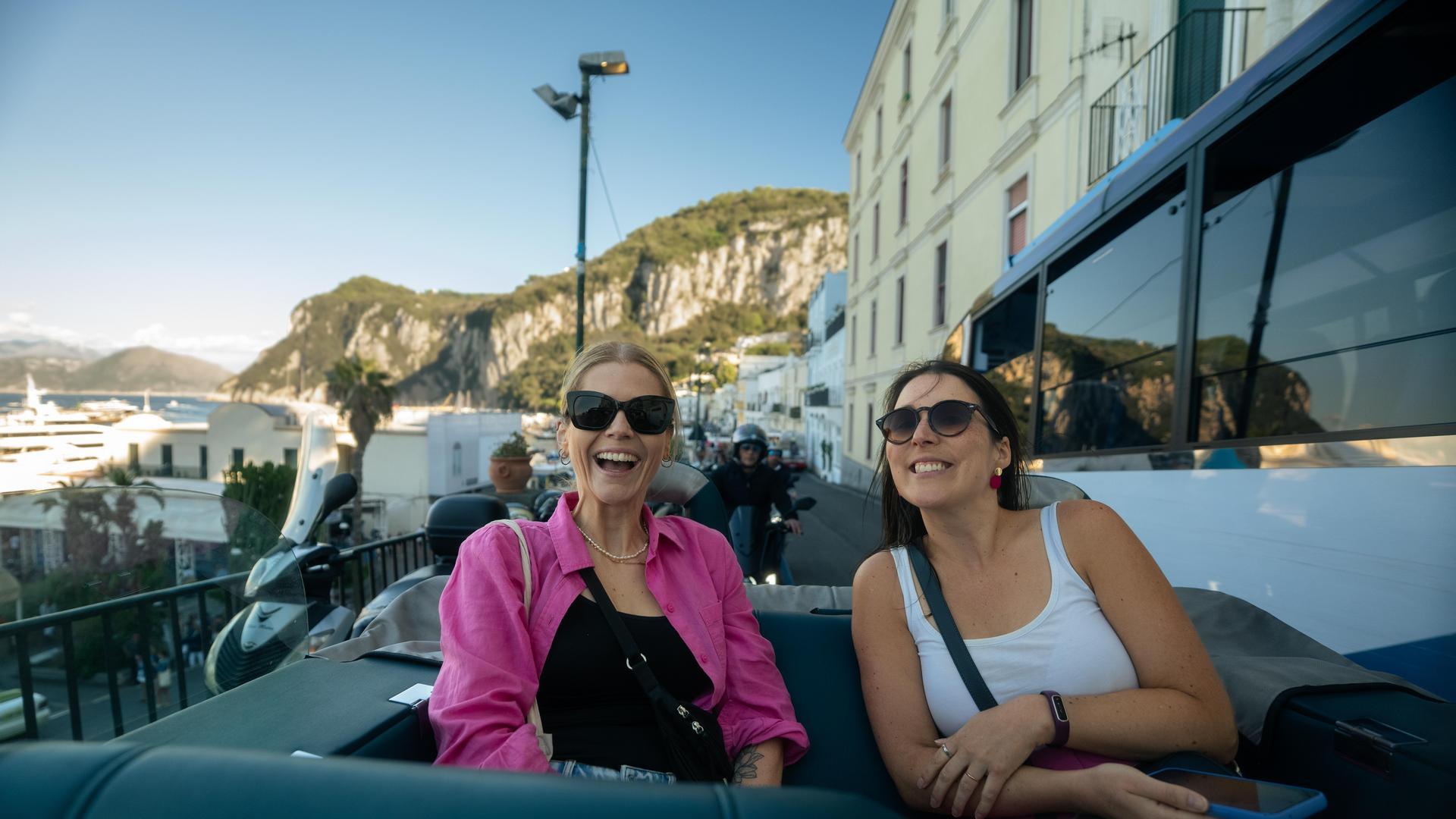 Two smiling women in sunglasses riding in the back of a convertible through the streets of Capri, with the island's rocky cliffs and marina visible in the background.
