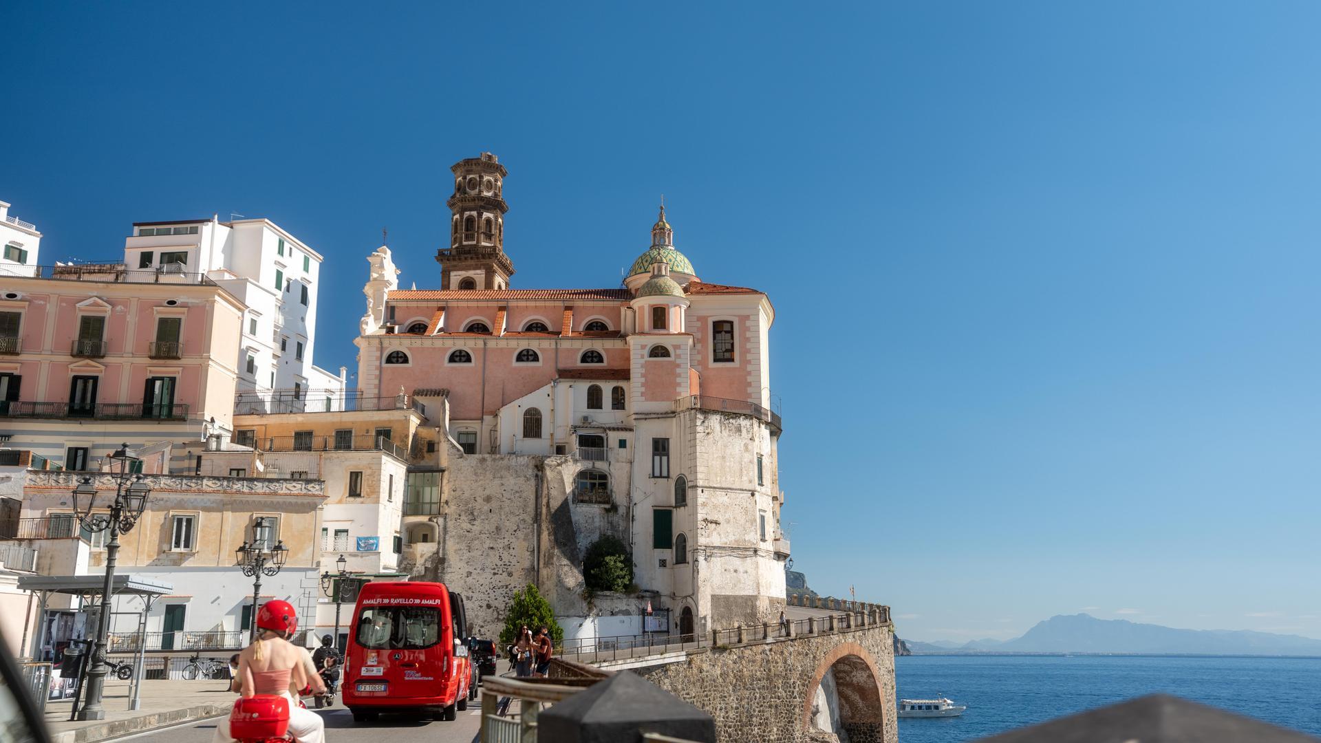 needs to be 1 concise sentence10:38Claude responded: Driving along the coastal road in Atrani on the Amalfi Coast, with a red van and scooter passing beneath the historic Collegiata di Santa Maria Maddalena churc…Driving along the coastal road in Atrani on the Amalfi Coast, with a red van and scooter passing beneath the historic Collegiata di Santa Maria Maddalena church.