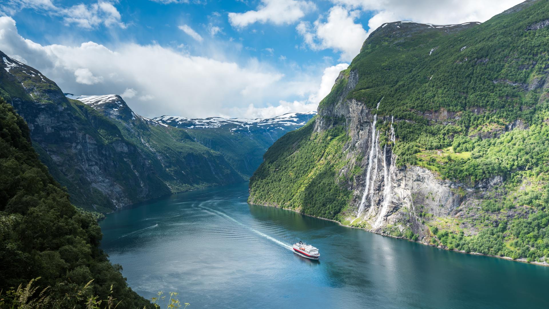 ship sailing along fjord with steep mountains and waterfall in background