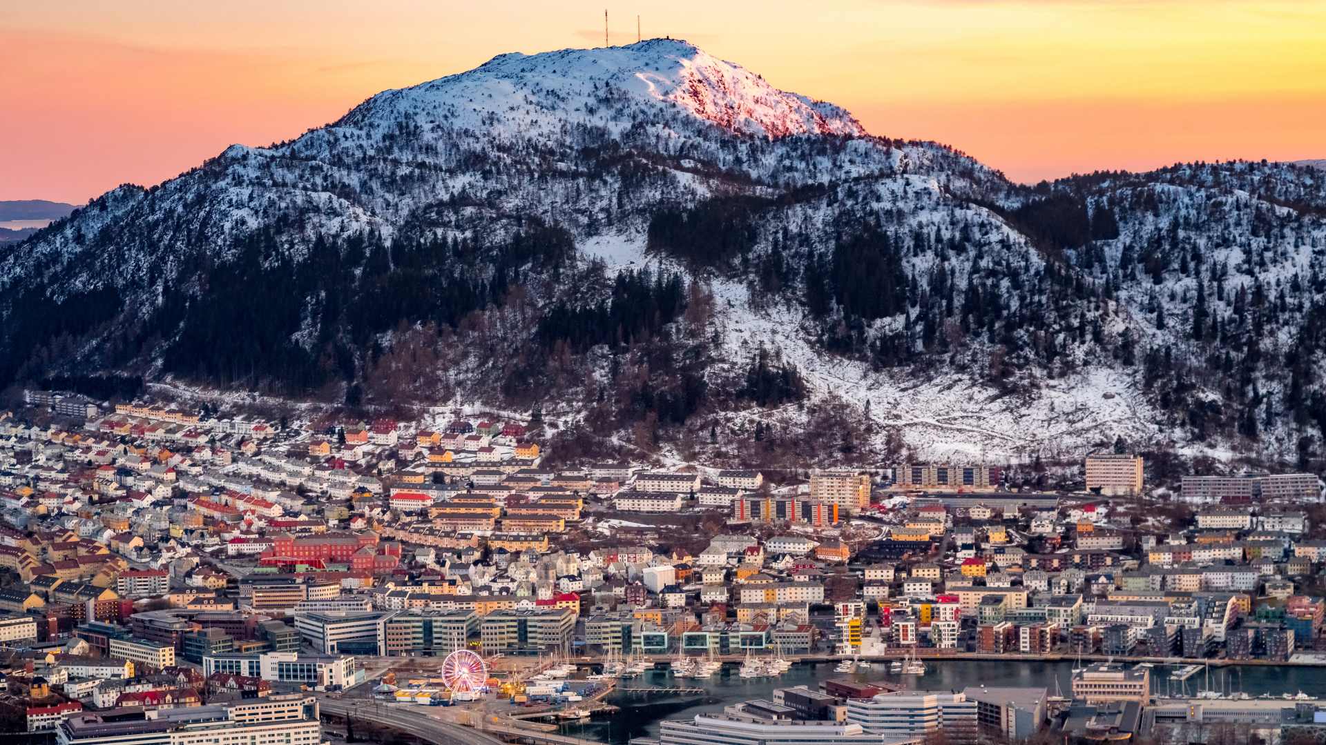Mount Lovstakken dusted in snow, overlooking the city of Bergen durtng an orange sunset.