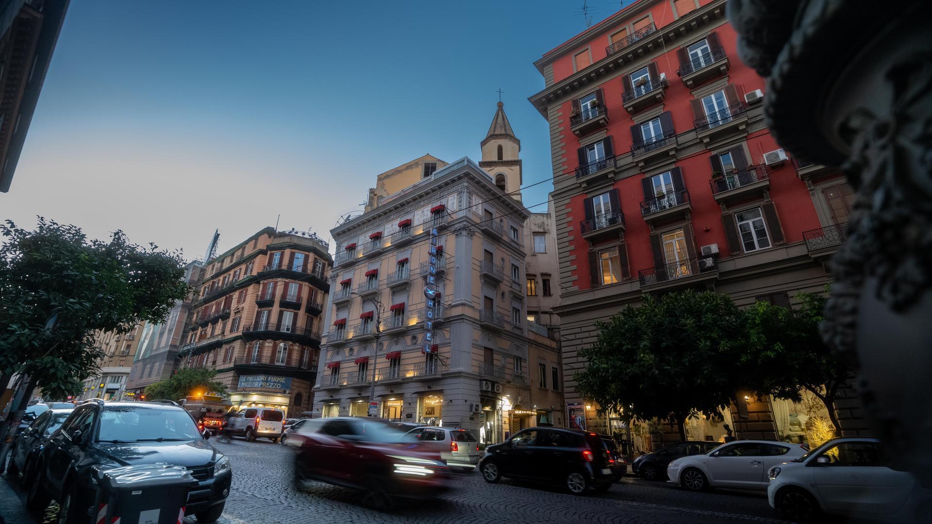 Busy evening traffic on a cobblestone street in Naples, surrounded by ornate multi-storey buildings and a lit hotel sign.