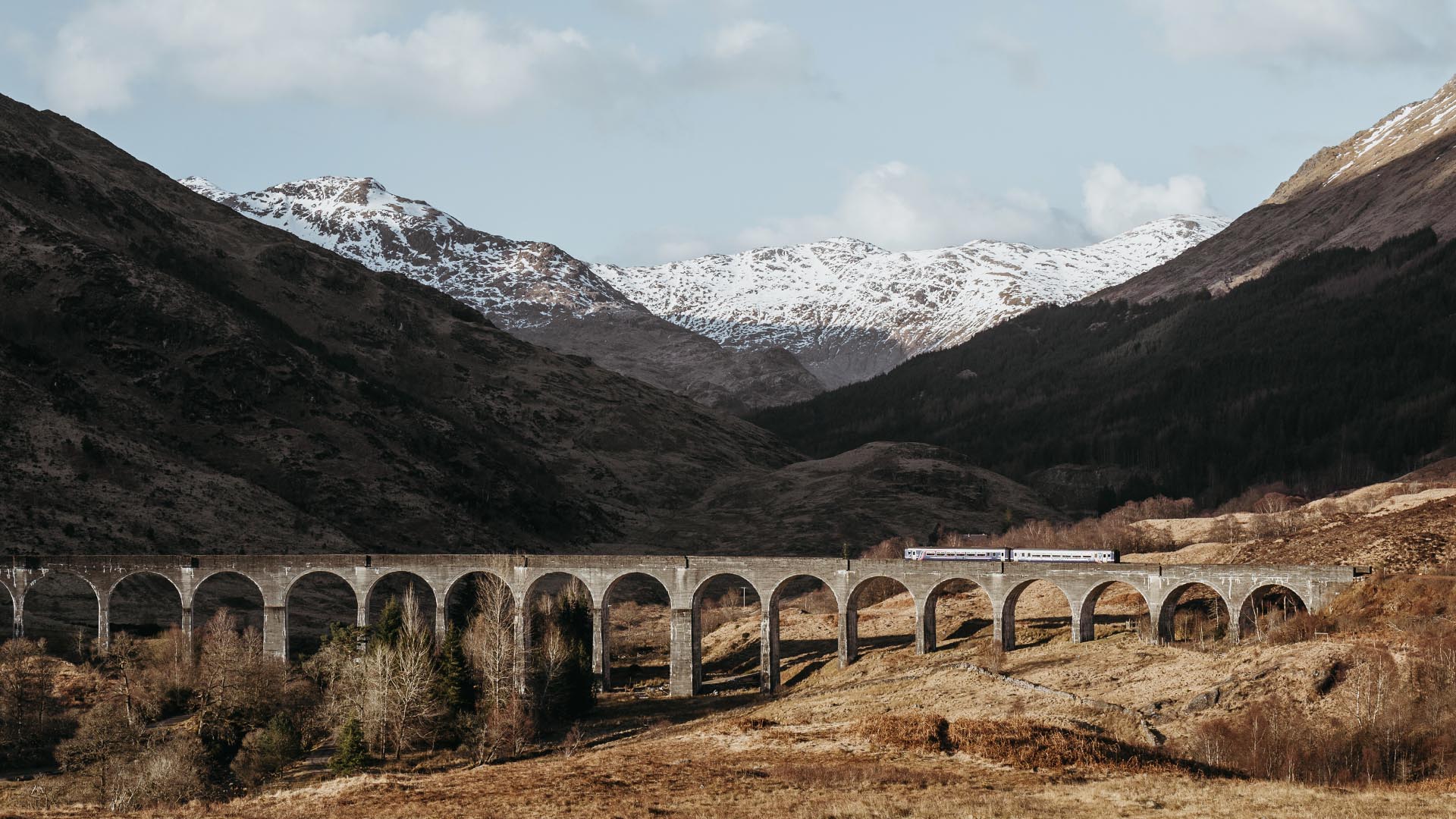 Glenfinnan Viaduct in Scotland with scot capped mountains behind the train passing over the bridge